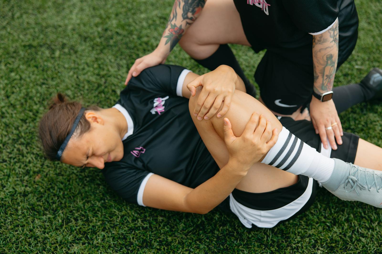 A female soccer player lying on grass, holding her injured knee, supported by a teammate.